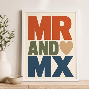 May include: A framed wall art print featuring the text MR in orange, AND in olive green, and MX in dark blue, with a tan heart symbol replacing the letter O. The design is set against a cream background, presented in a simple white frame.