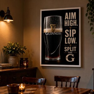 May include: A framed Guinness beer advertisement with a black background and white text that reads "AIM HIGH. SIP LOW. SPLIT THE G." The image features a pint of Guinness with the brand's logo. The poster is displayed in a room with a wooden table and chairs.
