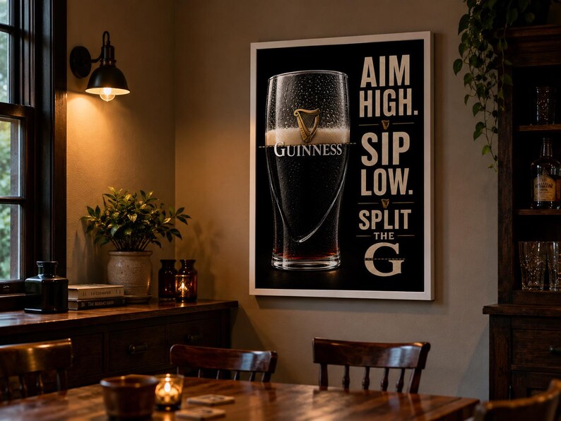 May include: A framed Guinness beer advertisement with a black background and white text that reads "AIM HIGH. SIP LOW. SPLIT THE G." The image features a pint of Guinness with the brand's logo. The poster is displayed in a room with a wooden table and chairs.