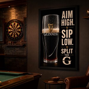 May include: A framed Guinness advertisement featuring a pint of stout with condensation. The black background has white text that reads "Aim High. Sip Low. Split the G." The scene includes a dartboard, pool table, and a leather armchair, suggesting a pub setting.