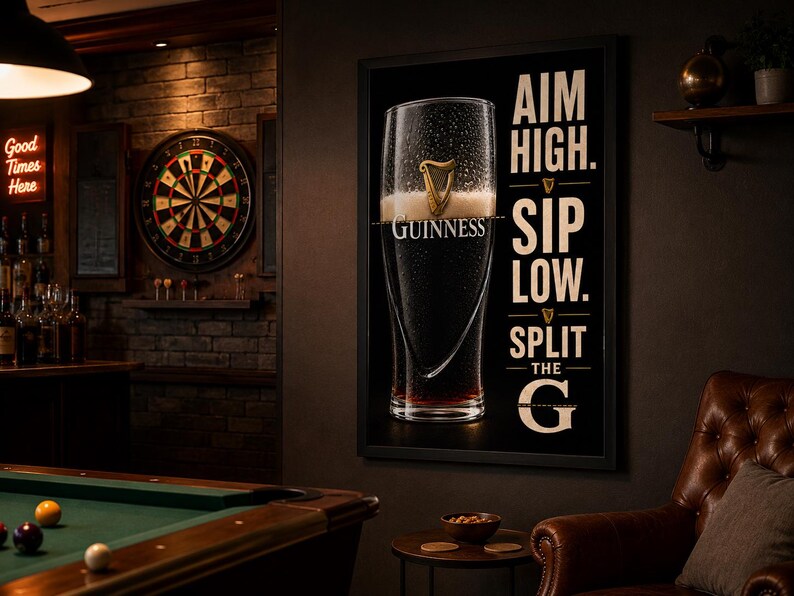 May include: A framed Guinness advertisement featuring a pint of stout with condensation. The black background has white text that reads "Aim High. Sip Low. Split the G." The scene includes a dartboard, pool table, and a leather armchair, suggesting a pub setting.
