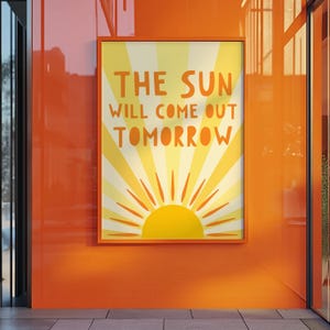 May include: A rectangular framed wall art print featuring a yellow sun graphic with radiating light beams against a pale yellow background. The text THE SUN WILL COME OUT TOMORROW is printed in bold orange letters across the center of the poster.