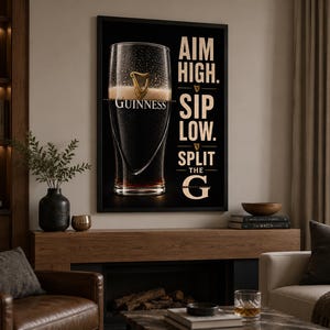 May include: A framed black and white Guinness advertisement. The image features a pint of Guinness with the text "AIM HIGH. SIP LOW. SPLIT THE G." The artwork is displayed in a living room setting with a fireplace and leather armchair.