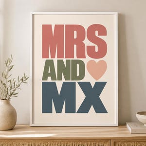 May include: A framed wall art print featuring the text MRS in large coral letters, AND in olive green letters, and MX in dark blue letters. A small, light pink heart icon is positioned to the right of the word AND, all set against a cream background.