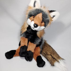 May include: A plush fox toy featuring a mix of brown, black, and white faux fur. The fox has pointed ears with white tips, amber-coloured eyes, a black nose, and a long, bushy tail with a white tip. The paws and chest area are solid black.