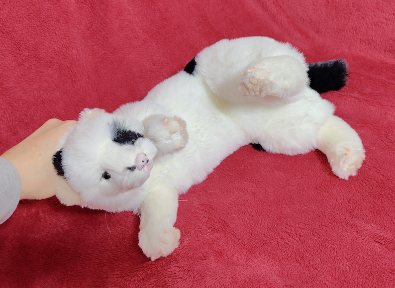 May include: A plush white cat toy with black spots on its head and back, and a black tail. The toy is lying on its back on a red surface. The cat toy is soft and fluffy.