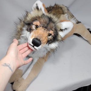 May include: A plush wolf toy featuring realistic brown, grey, and white faux fur. The stuffed animal has amber-coloured eyes, a black nose, and pointed ears with white inner fur. It is depicted in a lying position with extended front paws.