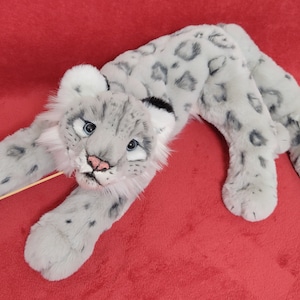 May include: A plush snow leopard toy with gray and white fur, lying on a red surface. The toy has a realistic appearance with black spots and a pink nose.