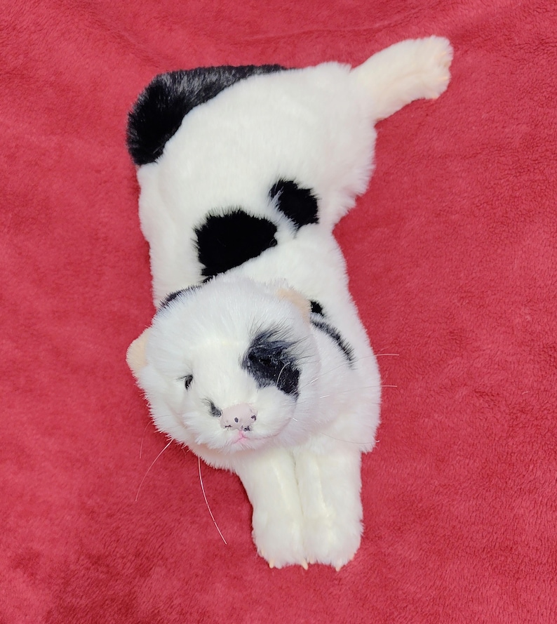 May include: A plush toy cat in a lying position, featuring a white body with black spots and markings. The cat has a pink nose and whiskers, with a black patch around one eye. The toy is set against a red background.