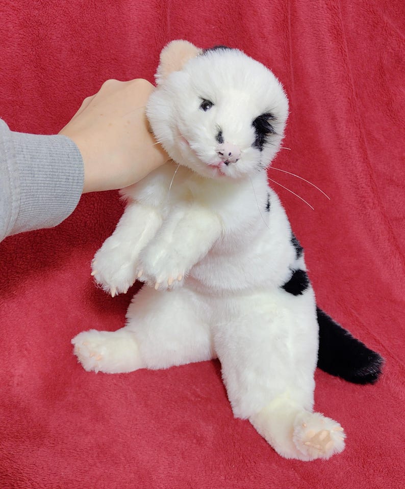 May include: A plush white cat with black spots and a black tail. The cat has a pink nose and tongue, and a black spot over one eye. The cat is sitting upright on a red surface.