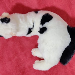 May include: A plush toy cat in a lying position, featuring a white body with black spots and a black tail. The toy is made of soft, fluffy material and is set against a red background.