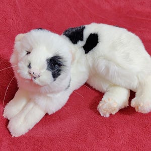 May include: A plush white cat with black spots on its head and back, lying on a red surface. The cat has a pink nose and whiskers, and its paws are visible. The toy is soft and fluffy.