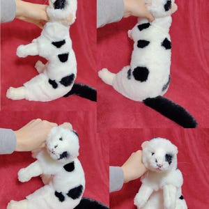 May include: A plush toy cat with a white body and black spots. The cat has a black tail and is being held up by a person. The toy cat has a cute, friendly face.