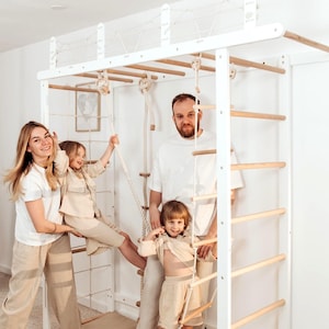 Puede incluir: Una estructura de escalada interior de madera blanca con escalera, barras de mono, escalera de cuerda y red de escalada. La estructura tiene peldaños de madera natural, se asienta sobre una alfombra beige y cuenta con un asiento de columpio.