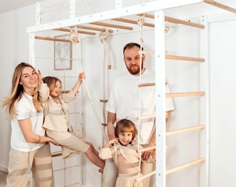 May include: A white wooden climbing frame with a ladder, rope, and rings. The frame is set up in a room with a beige mat on the floor. A family is interacting with the climbing frame.