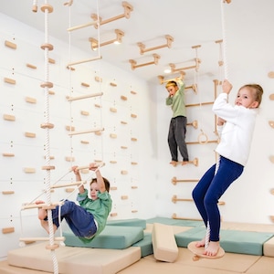 May include: A wooden indoor playground set featuring a climbing wall with light wood holds, a rope ladder, a hanging swing disc, and horizontal monkey bars. The structure is mounted to the wall and ceiling, complemented by soft beige and green floor mats.
