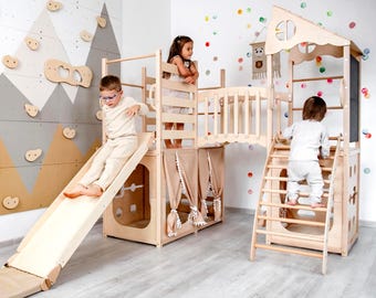 May include: A wooden indoor playground structure featuring a slide, a climbing ladder, a bridge, and a small playhouse area with beige curtains. The unit is constructed from light-colored wood and includes a flat mat at the base of the slide.