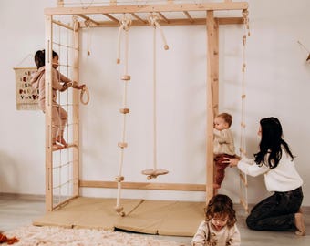 May include: A large wooden indoor jungle gym structure featuring a climbing net, monkey bars, a rope ladder, a hanging swing, and a rope climbing pole. The unit is constructed from light-coloured wood and includes a beige floor mat underneath.