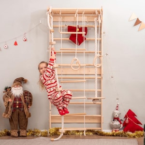 May include: A wooden climbing frame with a rope ladder, rings, and a child in Christmas pajamas. The frame is mounted on a wall, with a red bow and Christmas decorations. The child is climbing the rope ladder.