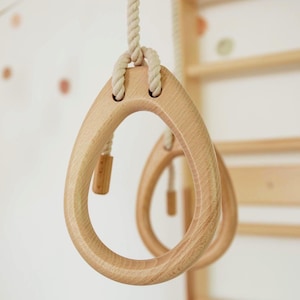 May include: A pair of light-colored wooden gymnastic rings with a smooth, rounded teardrop shape. Each ring is suspended by a thick, off-white twisted cotton rope that threads through two holes at the top of the wooden frame.