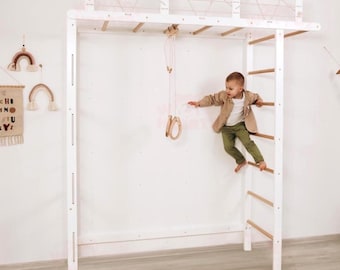 May include: A white wooden climbing frame with a ladder, horizontal bars, and a rope bridge. The frame includes hanging rings. A child is climbing the ladder. Decorative rainbows and a banner are on the wall.
