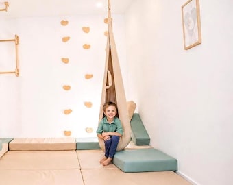 Può includere: Un'altalena a nido in tessuto beige sospesa al soffitto, posizionata sopra una serie di tappetini da pavimento color beige e verde salvia. La struttura include una parete da arrampicata con prese in legno e un quadro appeso alla parete.
