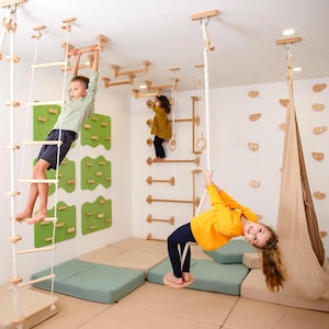 May include: Indoor playground with wooden climbing structures, including a rope ladder, horizontal bars, and climbing holds. Children are shown playing on the equipment. The room features green and beige mats and a beige swing.