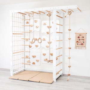 May include: A white wooden indoor jungle gym featuring a climbing net, a rope ladder, gymnastic rings, a climbing wall with wooden holds, and a hanging rope with wooden blocks. A beige floor mat sits below, and an alphabet wall hanging is nearby.
