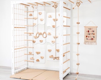 May include: A white wooden climbing frame with various features, including a rope ladder, climbing holds, gymnastic rings, and a hanging rope ladder. The frame has a beige mat at the base and a decorative alphabet banner.