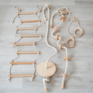Puede incluir: Un conjunto de juguetes de escalada para niños hechos de madera natural y cuerda blanquecina. El conjunto incluye una escalera de cuerda, una cuerda de escalada con discos de madera, un columpio y anillas de gimnasia.