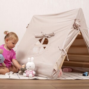 May include: A beige canvas play tent with a wooden frame, featuring a circular window and tie closures. A young child in a pink shirt sits on a rug with stuffed animals, including a bear and a bunny, creating a cozy play area.