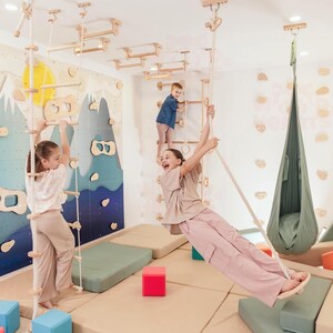 May include: Indoor wooden playground with climbing structures, ladders, and a swing. Children are playing on the equipment. The room has a colorful mural and soft mats on the floor. The equipment is made of light-colored wood.