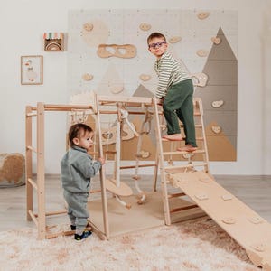 May include: A wooden indoor jungle gym climbing structure featuring a ladder, a slide with climbing holds, a rope net, and hanging rope accessories. The light-colored wood frame is designed for active play and includes a flat base platform.