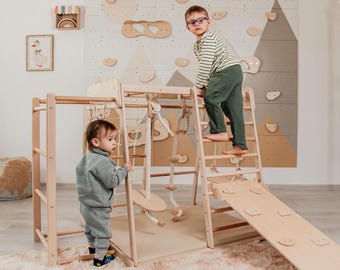 May include: A wooden climbing frame with a ladder, slide, and rope swing. Two children are playing on the frame. The background features a wall with climbing holds and a mountain mural. A rug is on the floor.
