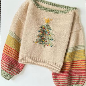 Könnte beinhalten: Ein beiger Strickpullover mit grünem Ausschnitt und gestreiften Ärmeln. Der Pullover zeigt einen gestickten Weihnachtsbaum mit bunten Ornamenten.