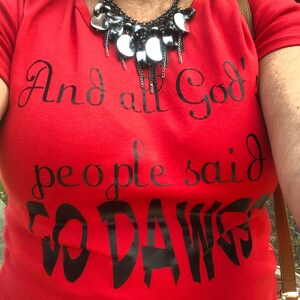 May include: A red t-shirt with the text "And all God's people said GO DAWGS" in black script. A black and silver necklace is worn over the shirt.