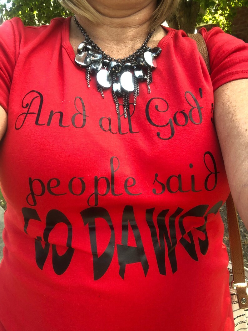 May include: A red t-shirt with the text "And all God's people said GO DAWGS" in black script. A black and silver necklace is worn over the shirt.