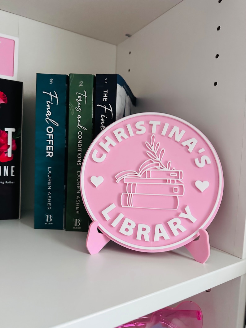 May include: Pink round sign with white lettering that says "Christina's Library". The sign has a stack of books, a heart, and a leaf design. The sign is on a white shelf with books behind it.