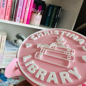 May include: A pink round sign with white lettering that reads "Christina Library". The sign has a white heart and a stack of books with leaves on top.