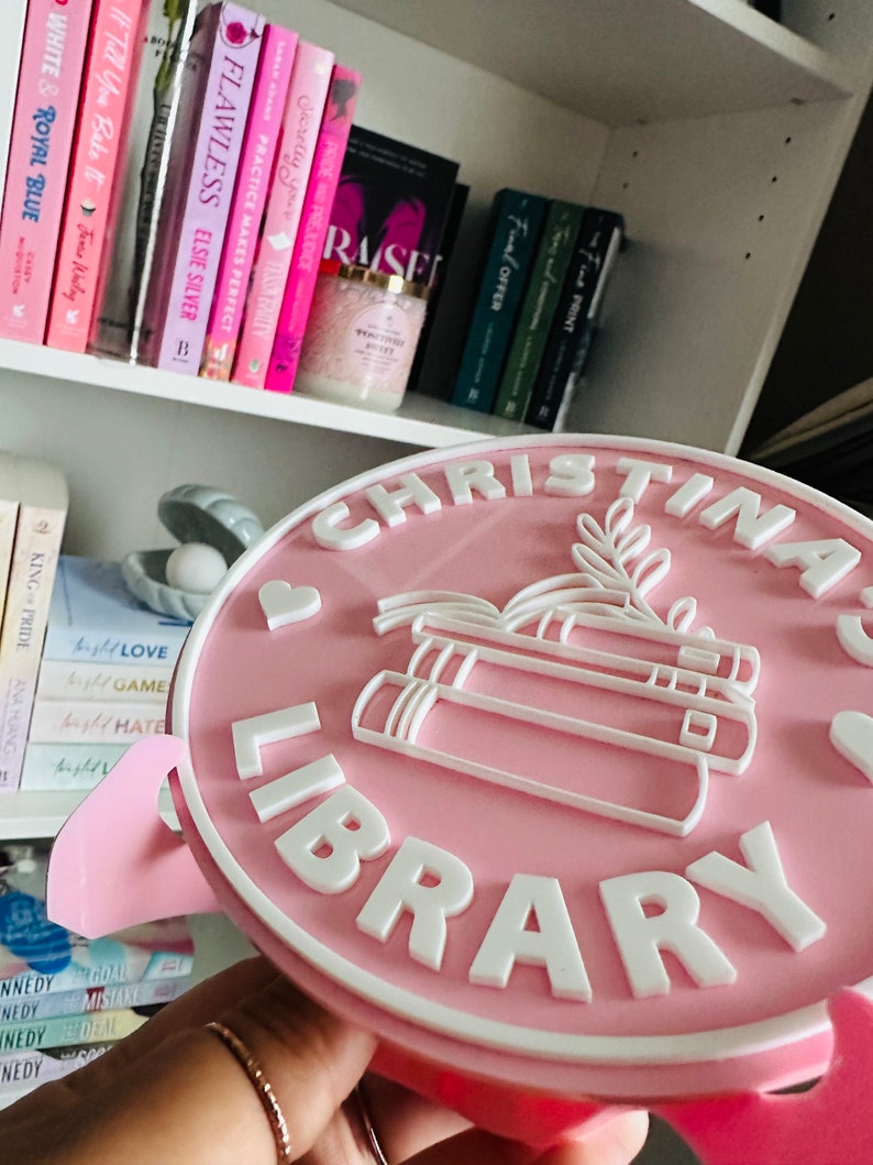 May include: A pink round sign with white lettering that reads "Christina Library". The sign has a white heart and a stack of books with leaves on top.