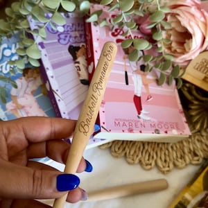 May include: A wooden baseball bat with the words "Baseball Romance Era" engraved on it. The bat is held in a hand with blue nail polish. The bat is in front of a stack of books with pink and purple covers.