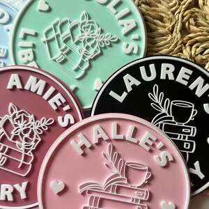 May include: Four pink, green, black, and white round coasters with white text that reads "Julia's Library", "Halle's Library", "Lauren's Library", and "Amie's Library". Each coaster features a white illustration of a stack of books with a cup of tea and a flower.
