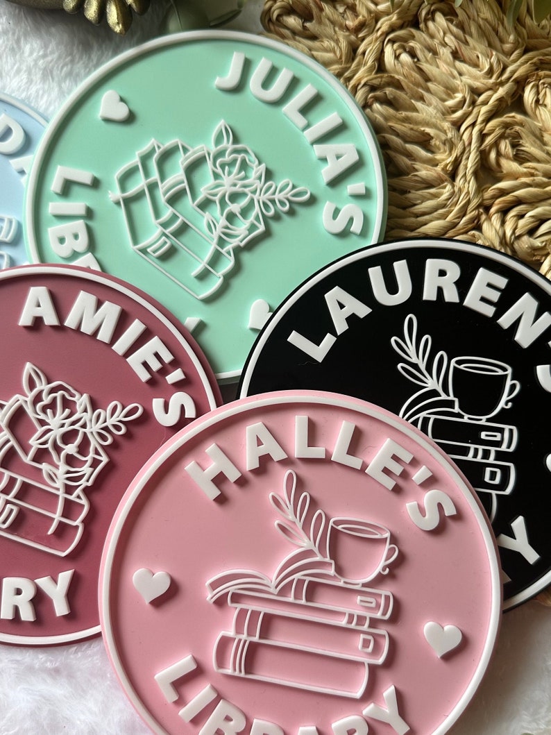 May include: Four pink, green, black, and white round coasters with white text that reads "Julia's Library", "Halle's Library", "Lauren's Library", and "Amie's Library". Each coaster features a white illustration of a stack of books with a cup of tea and a flower.