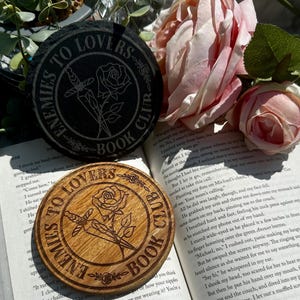 May include: Two coasters with a rose and dagger design. One coaster is made of black slate and the other is made of wood. Both coasters have the text "Enemies to Lovers Book Club".