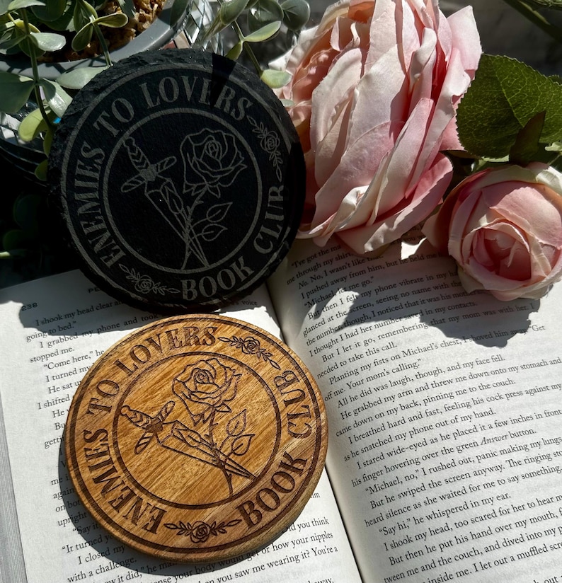 May include: Two coasters with a rose and dagger design. One coaster is made of black slate and the other is made of wood. Both coasters have the text "Enemies to Lovers Book Club".