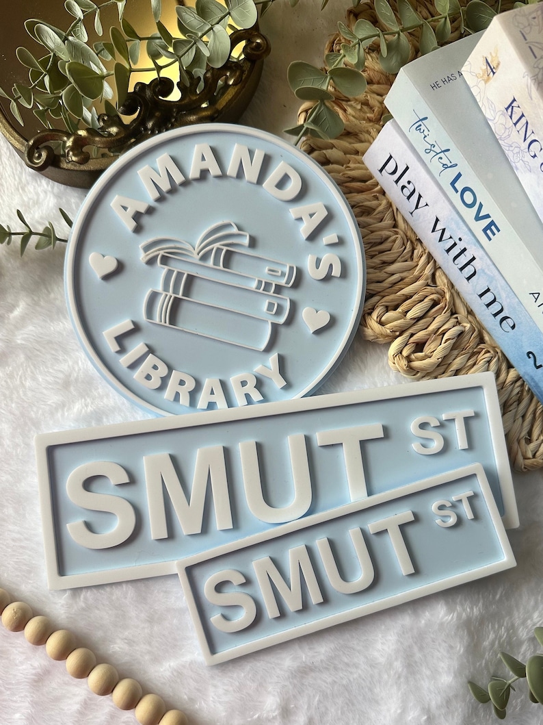 May include: A blue and white round sign with the text "Amanda's Library" and a stack of books with a heart on each side. Two blue and white rectangular signs with the text "SMUT ST" in white.