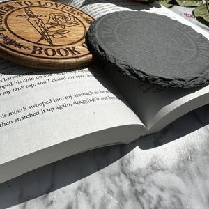 May include: A wooden coaster with the text "Thieves to Lovers Book Club" and a rose and dagger design. The coaster is sitting on top of a book with a slate coaster on top of the book.