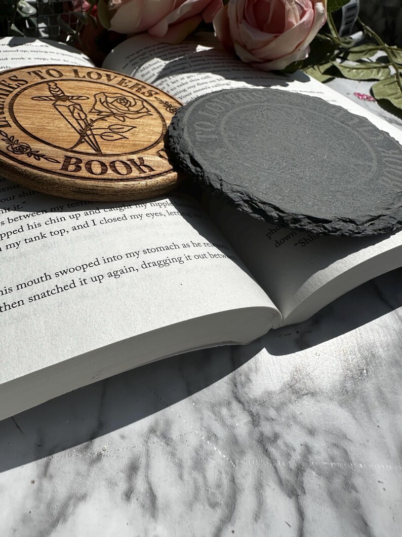 May include: A wooden coaster with the text "Thieves to Lovers Book Club" and a rose and dagger design. The coaster is sitting on top of a book with a slate coaster on top of the book.