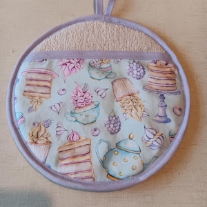 May include: A round, light blue and purple pot holder with a white terry cloth backing. The pot holder features a pattern of teacups, cakes, and teapots with a light blue background.
