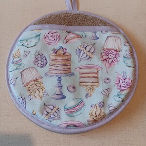 May include: A round, light blue pot holder with a light purple trim and a brown terry cloth back. The front features a pattern of cakes, cupcakes, macarons, and teacups in pastel colours.